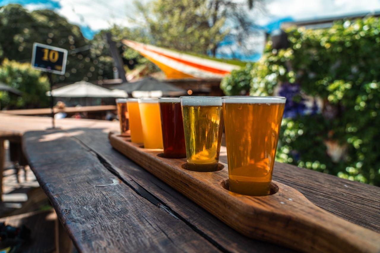 gallery-5 A vibrant beer flight displayed on a rustic wood table outdoors during a sunny day.